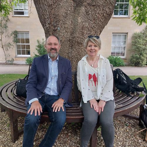 Prof Andrew Rice and BBC Radio presenter Fiona Talkington sitting on a bench under a tree, facing the camera