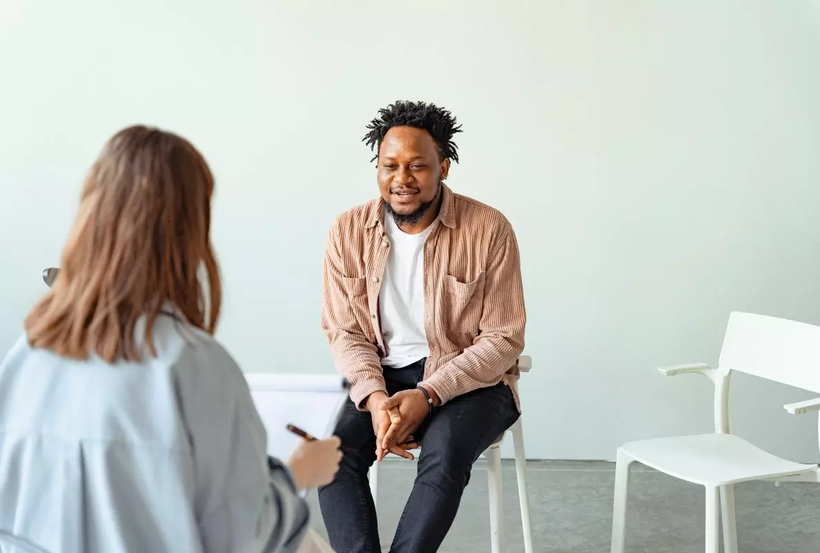 A man is sitting on a chair, talking to a woman holding a clipboard with her back to the camera
