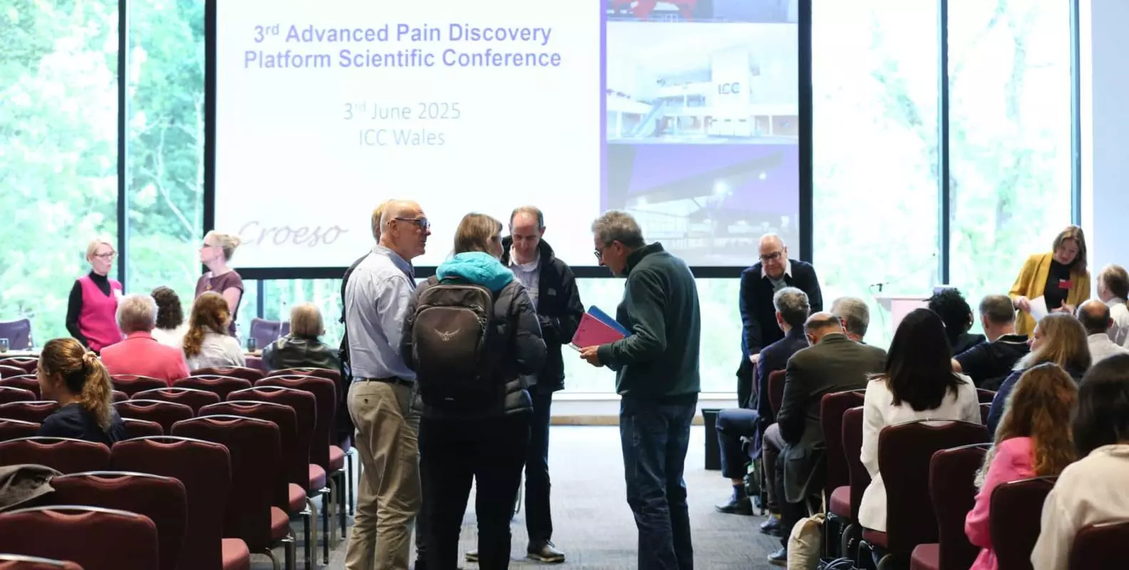 Photo of a display of slides that read "3rd APDP conference, June 2025" in the background. There are people sitting and standing in a group in the foreground.
